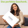 Strahlende Frau hält eine stilvolle weiße Box von feela, betitelt als 'Das perfekte Geschenk', ideal für jemanden, der Komfort und Design schätzt.