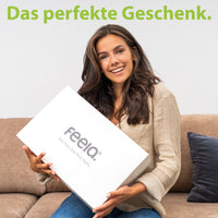 Strahlende Frau hält eine stilvolle weiße Box von feela, betitelt als 'Das perfekte Geschenk', ideal für jemanden, der Komfort und Design schätzt.
